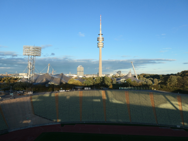 Olympiastadion in Berlin mit dem Fernsehturm im Hintergrund, umgeben von B"umen, Geb"uden und Lichtern unter einem bew"olkten Himmel.