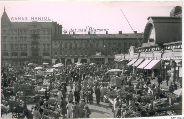 Schwarz-weiß-Foto eines belebten Berliner Markts mit Menschen, Gemüsewagen und Gebäuden mit Schildern im Hintergrund.
