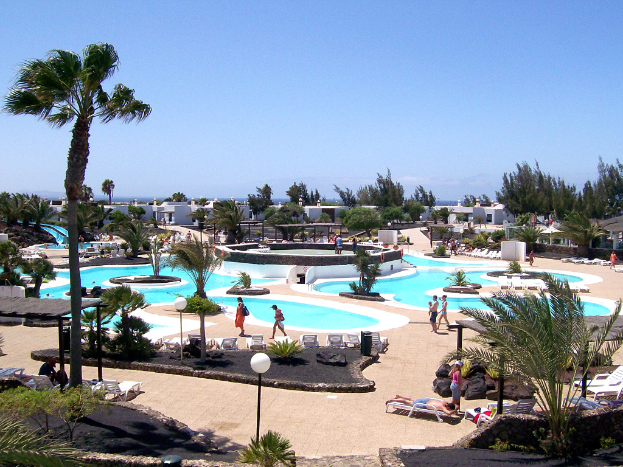 Ein Resort mit von Bäumen umgebenen Pools, Menschen, die dazwischen gehen, Gebäuden im Hintergrund und einem klaren Himmel darüber.