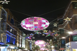 Eine festliche Stadtstraße bei Nacht mit zahlreichen Lichtern und Dekorationen, darunter eine große Diskokugel, die von einem Gebäude hängt.