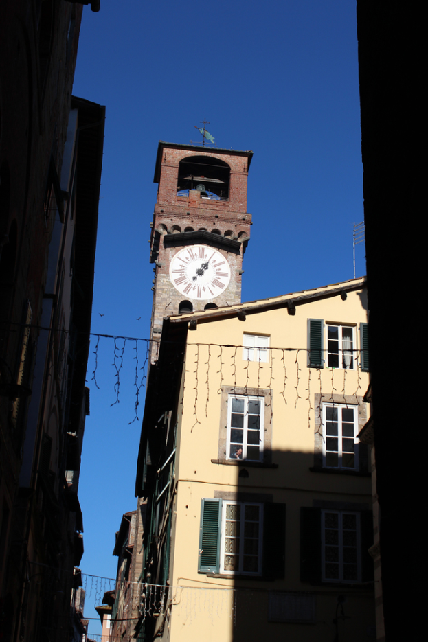 Ein Uhrenturm mit einer Antenne oben, umgeben von Gebäuden mit Glasfenstern, unter einem blauen Himmel.