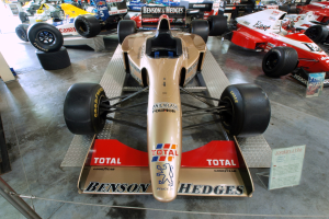 Eine Gruppe von Rennwagen in einem Motorsportmuseum, getrennt durch einen Metallzaun mit einer Texttafel links und Menschen, Lichter und andere Ausstellungsstücke im Hintergrund.