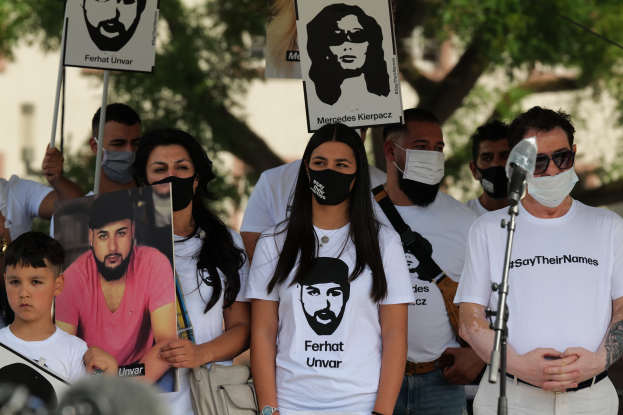 Eine Gruppe von Menschen mit Masken und Schildern auf einer Demonstration in Mexiko-Stadt, mit einem Mikrofon und einer Kamera im Vordergrund und Bäumen und Gebäuden im Hintergrund.
