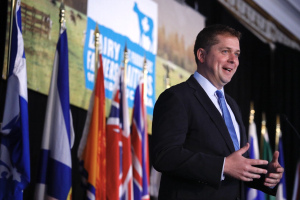 Mann in Anzug und Krawatte, der an einem Rednerpult mit Mikrofonen steht, umgeben von Fahnen und einem Bildschirm im Hintergrund.