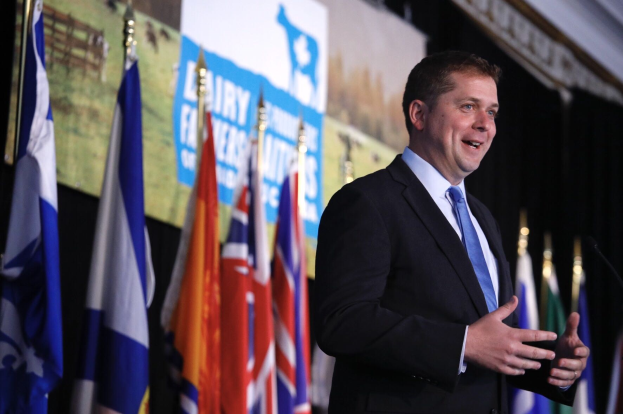 Mann in Anzug und Krawatte, der an einem Rednerpult mit Mikrofonen steht, umgeben von Fahnen und einem Bildschirm im Hintergrund.