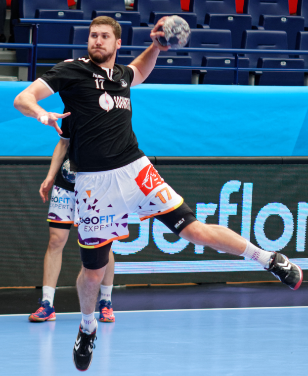 Ein Mann in schwarzem Hemd und weißen Shorts hält einen Handball und bereitet sich auf ein Spiel in einem Stadion mit Zuschauern im Hintergrund vor.