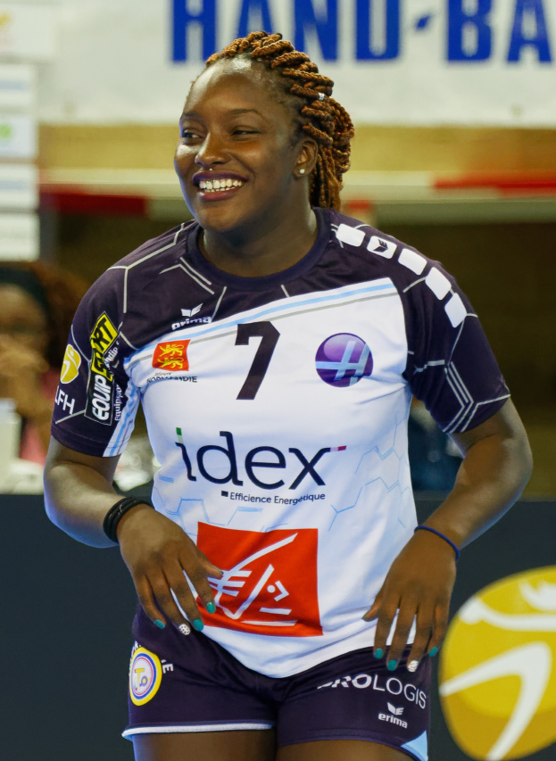 Eine Frau in einem lilafarbenen und weißen Trikot steht selbstbewusst auf einem Handballfeld, lächelt und hat ein "FIVB FIVB Handball World Cup 2019"-Schild im Hintergrund.