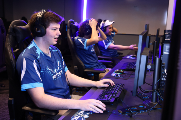 Gruppe junger Männer mit Headsets, die ein Videospiel auf Computern mit Monitoren, Tastaturen und Mäusen an einem Schreibtisch spielen, mit beleuchteter Wand im Hintergrund.