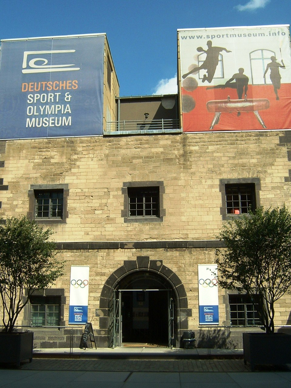 Außenansicht des Deutschen Sport & Olympiamuseums in Berlin, das ein Gebäude mit Fenstern und einer Tür zeigt, umgeben von Bäumen und Bannern, unter einem bewölkten Himmel.