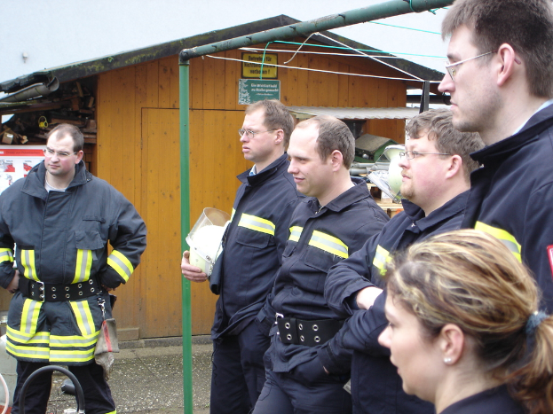 Eine Gruppe von Männern in blauen Feuerwehranzügen, die nach links schauen, mit einem kleinen hölzernen Schuppen und Rohrrahmen im Hintergrund.