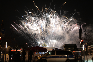 Ein Polizeiwagen vor einer Menschenmenge bei einer Silvesterfeier in Berlin, mit farbigen Feuerwerken am Himmel und Stadtlichtern im Hintergrund.