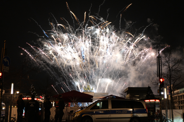 Ein Polizeiwagen vor einer Menschenmenge bei einer Silvesterfeier in Berlin, mit farbigen Feuerwerken am Himmel und Stadtlichtern im Hintergrund.