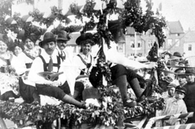 Eine Gruppe von Menschen in traditioneller bayrischer Tracht fährt auf einem mit Blättern geschmückten Wagen in einem Umzug, einige spielen Musikinstrumente, mit Gebäuden und einem klaren Himmel im Hintergrund, dargestellt in Schwarz-Weiß.