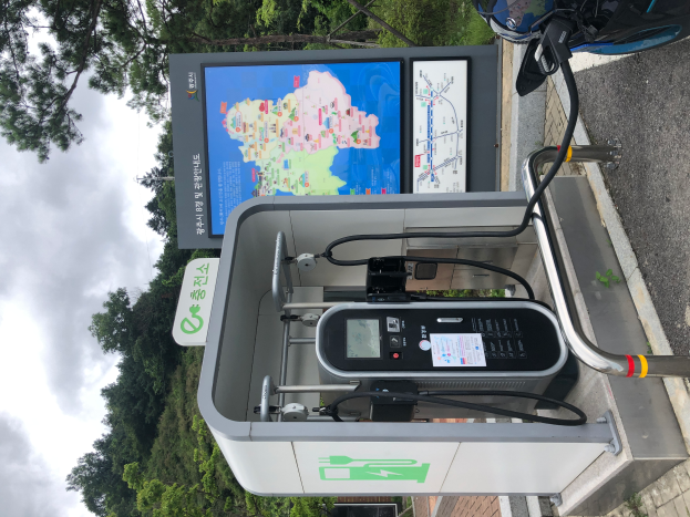 Eine Ladesäule für Elektrofahrzeuge am Straßenrand mit einem Motorrad in der Nähe, Bäumen und einem klaren blauen Himmel im Hintergrund und einem digitalen Anzeigebrett, das darauf hinweist, dass es sich um eine E-Bike-Ladestation handelt.