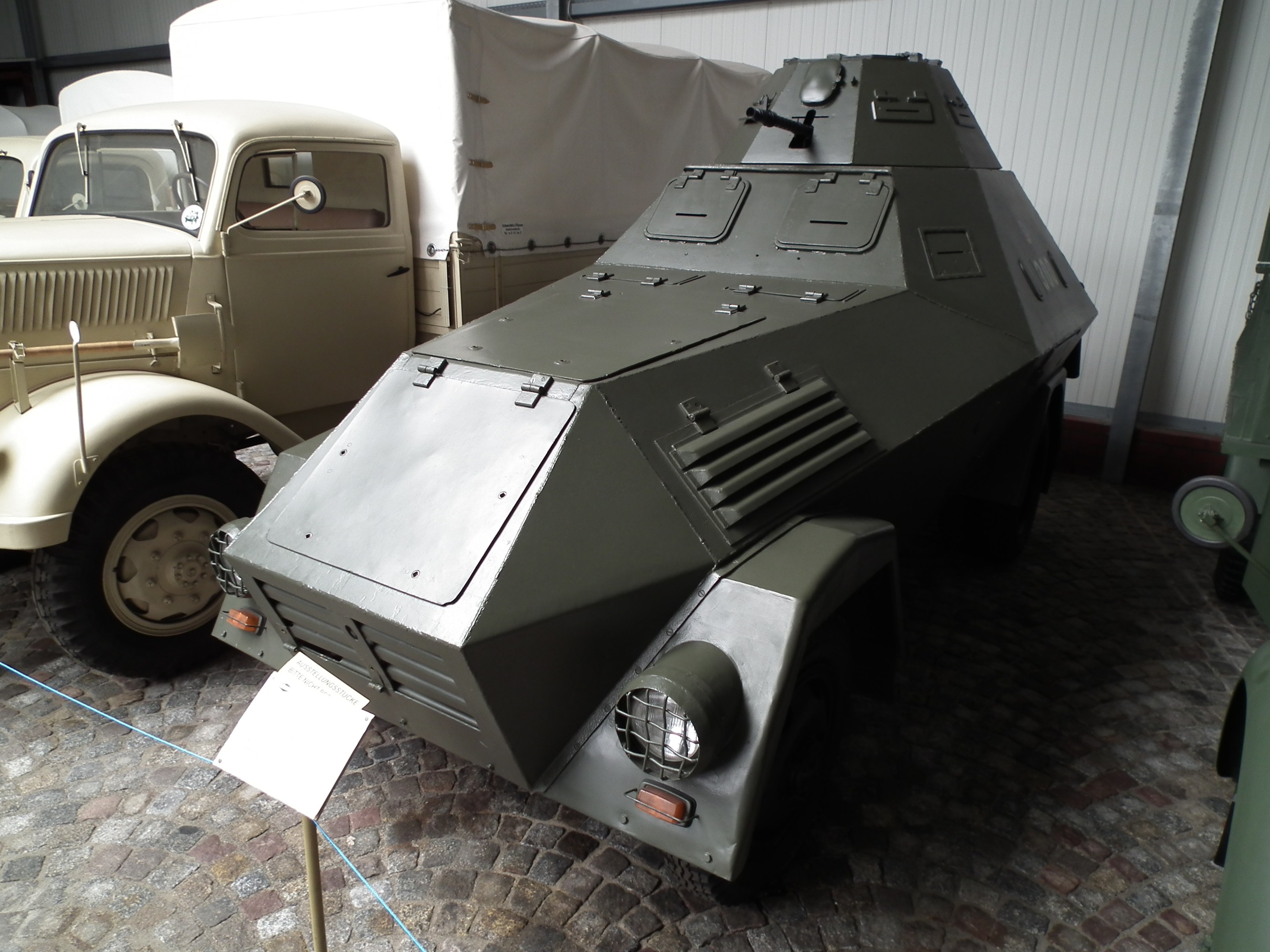 Panzerfahrzeug in einer Garage neben anderen Militärfahrzeugen geparkt, mit einer Tafel auf einem Ständer auf dem Boden und einer Wand im Hintergrund.