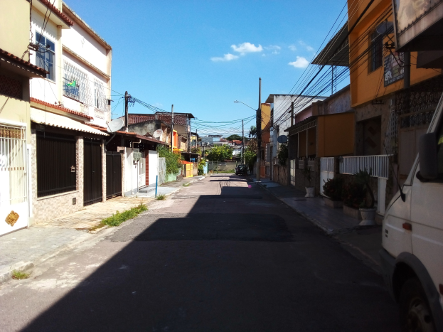 Leere Straße mit einem Van auf der Seite geparkt, flankiert von Gebäuden, Strommasten mit Drähten, Bäumen im Hintergrund und einem bewölkten Himmel.