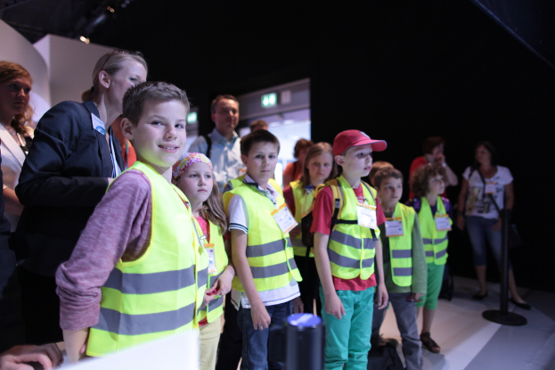 Eine Gruppe von Kindern in Sicherheitswesten und Namensschildern, mit einem Jungen auf der linken Seite, der lächelt, auf einem Boden mit einem Pfahl auf der rechten Seite und einigen Menschen im Hintergrund.