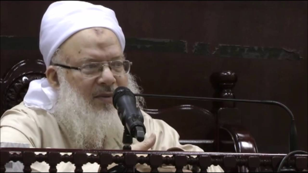 Asaduddin Owaisi, ein Mann mit einem weißen Bart und Turban, spricht in ein Mikrofon und trägt ein cremefarbenes Gewand und eine Brille, vor einem hölzernen Geländer auf einem dunklen Hintergrund.