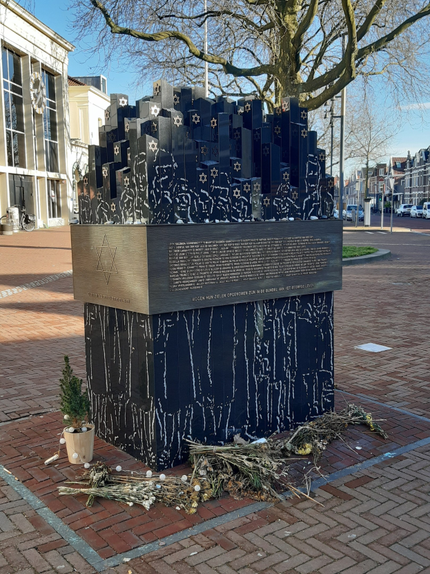 Denkmal in einem Stadtplatz für Holocaust-Opfer mit einem Baum davor, einem Topfpflanze auf dem Boden, verstreuten trockenen Blättern, umgeben von Gebäuden, Fahrzeugen, Masten, Bäumen und Himmel.