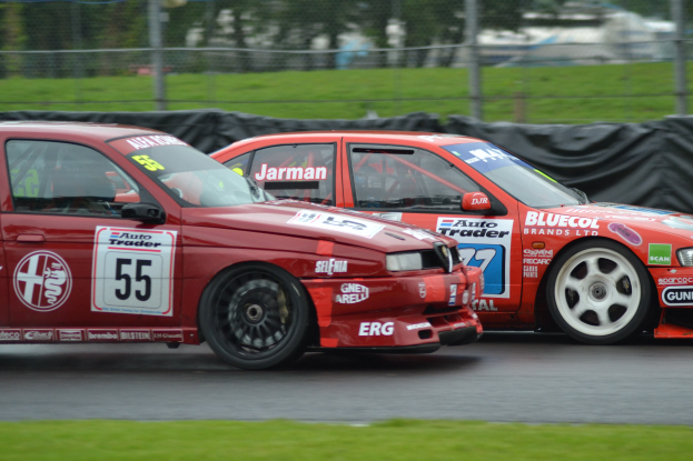 Zwei rote Rennwagen mit Nummern und Text auf ihnen, die auf einer Rennstrecke fahren, mit einem unscharfen Hintergrund aus einem Zaun, Bäumen und anderen Gegenständen.