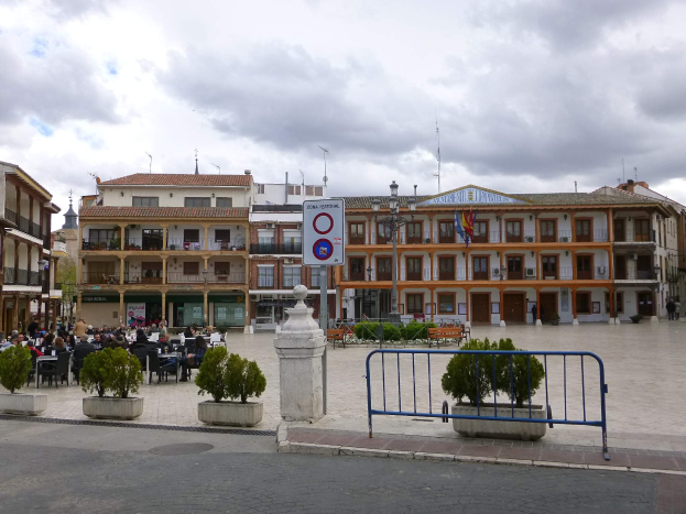 Ein belebter Stadtplatz mit Menschen auf Stühlen sitzend und stehend, umgeben von Topfpflanzen, Metallabsperrungen, Schildern, Straßenlaternen mit Flaggen, Gebäuden und einem bewölkten Himmel.