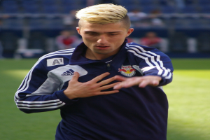 Ein junger Mann in einem blauen T-Shirt steht auf einem Fußballfeld, mit entschlossener Miene und ausgestreckten Armen, während der Hintergrund unscharf ist.