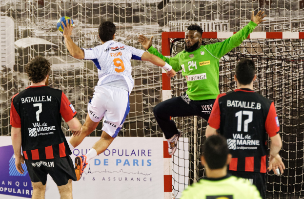 Eine Gruppe von Männern, die Handball spielen, mit einem, der springt und den Ball hält, vor einem Torpfosten und einer Tafel mit der Aufschrift "Ligue 1 - Bordeaux vs Marseille".