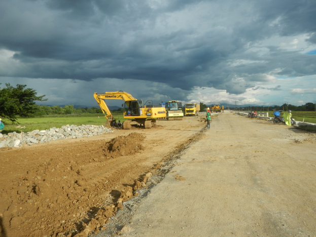 Gruppe von Bauarbeitern auf einer Schotterstraße mit Kränen, Steinen, Gras, Bäumen und Strommasten im Hintergrund unter einem bewölkten Himmel.