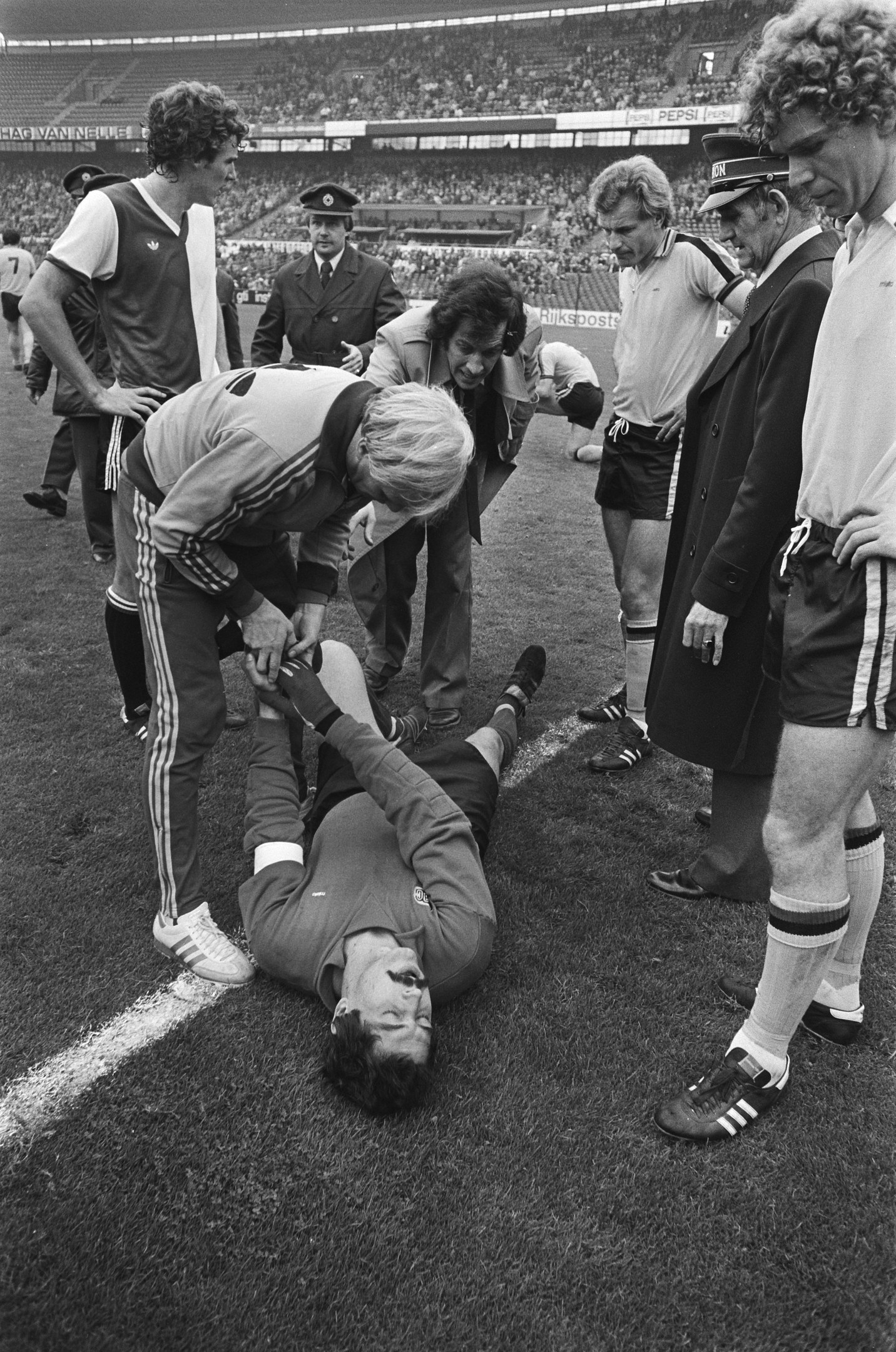 Schwarzes und weißes Bild eines Fußballspielers, der auf dem Boden liegt und von einer Gruppe Männer umgeben ist, mit einer Menschenmenge im Hintergrund der Stadion.