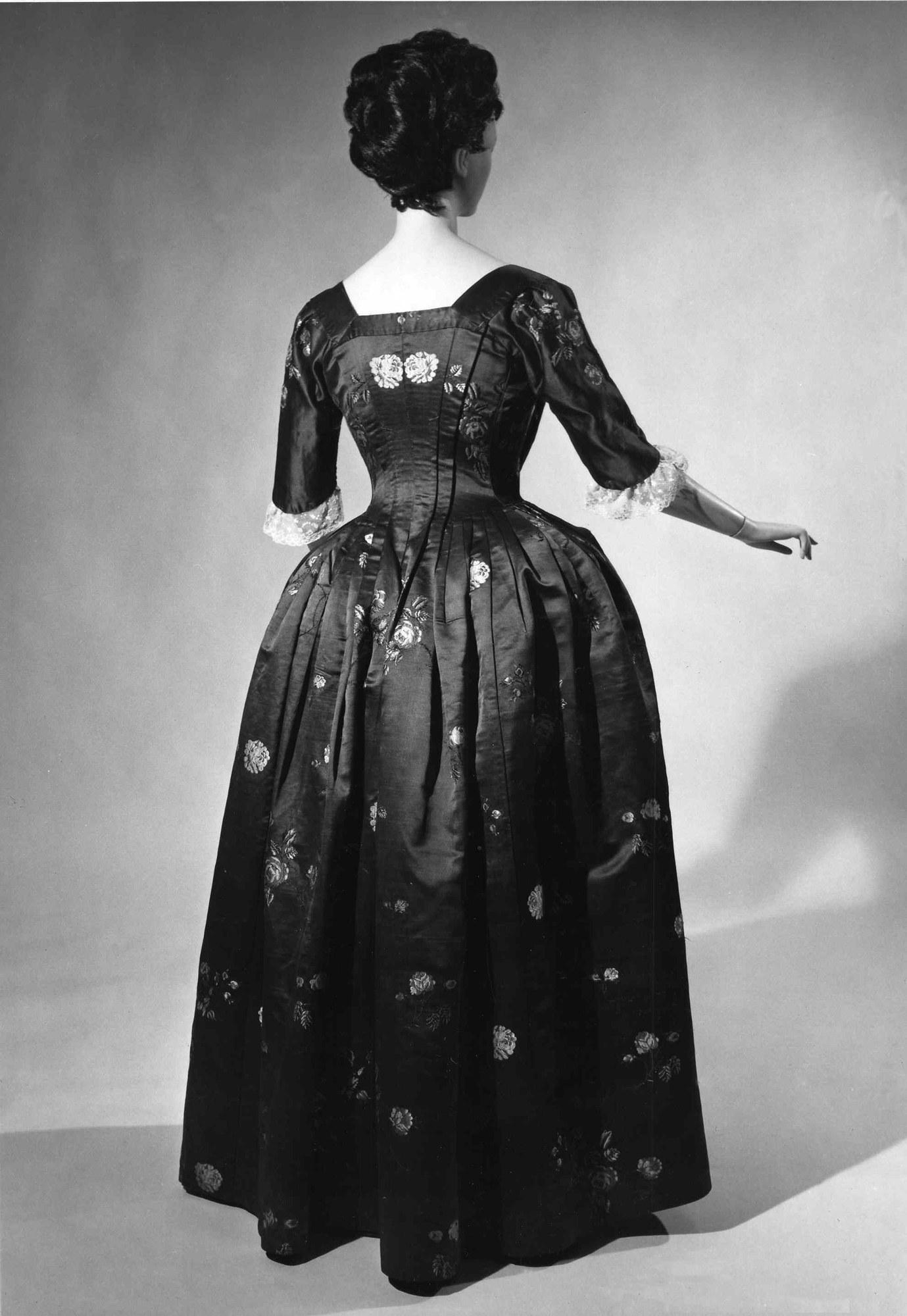 Eine Frau in einem 1950er-Kleid mit langem, engem Oberteil und vollem Rock posiert in einem Schwarz-Weiß-Foto, die Hände gefaltet, schwarze High Heels und ein klassisches Haar mit dezentem Make-up.