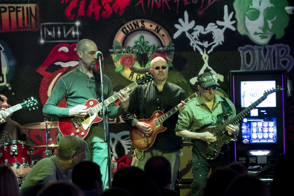 Drei Männer auf einer Bühne bei einem Konzert, mit einer Menge unten und einer gemalten Wand im Hintergrund.