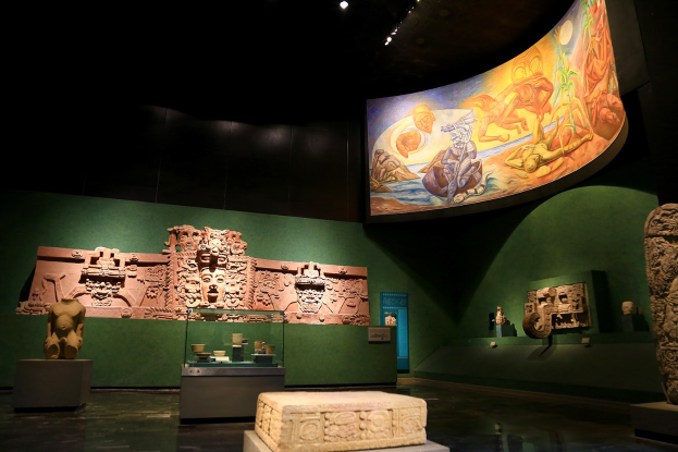 Ein Museumsraum mit verschiedenen Artefakten, darunter Skulpturen, Tischgegenstände, eine Texttafel, ein Wandgemälde und Deckenleuchten im Mexiko-Stadt-Museum der Anthropologie.