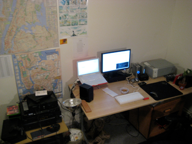 Ein Raum mit einem Tisch, auf dem ein Laptop, ein Lautsprecher, ein Monitor, eine Tastatur, ein Ball, eine Maus, ein Scanner und Papiere liegen, sowie ein weiterer Tisch mit elektronischen Geräten, mit Charts und Postern an der Wand dahinter.