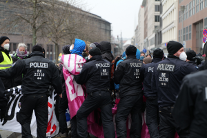 Eine Gruppe von Polizisten steht vor einer Menschenmenge, einige tragen Mützen und Masken, mit einem Banner im Vordergrund und Gebäuden, Bäumen und Schildern im Hintergrund unter einem klaren blauen Himmel.