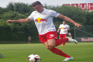 Ein professioneller Fußballspieler kickt einen Ball auf einem Rasenfeld mit "RB Leipzig" im Hintergrund bei einem klaren blauen Himmel.