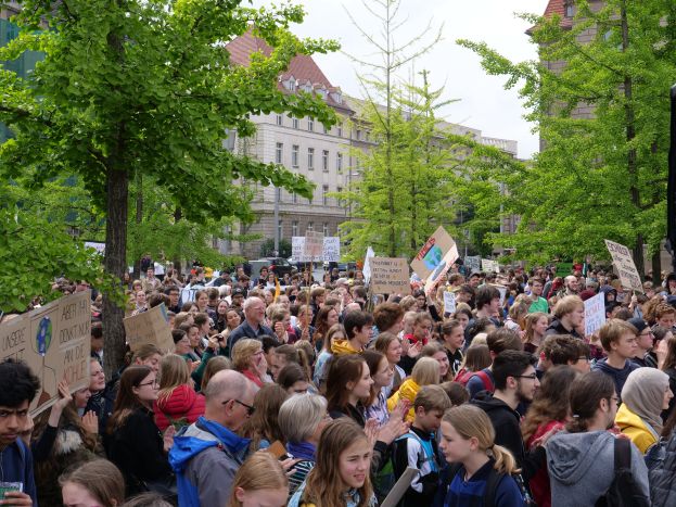 Eine große Menge Menschen mit Schildern steht vor einem Gebäude in Berlin, mit Bäumen, Fahrzeugen und einem Lautsprecher, unter einem klaren Himmel.