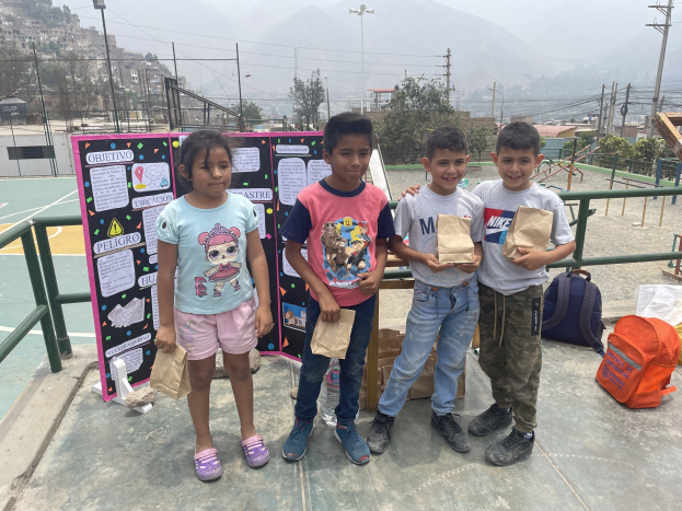 Gruppe von Kindern, die auf einem Basketballfeld stehen und Papiertüten halten, mit Vorräten auf dem Boden und einer Tafel mit Text links daneben, vor dem Hintergrund von Einzäunung, Masten, Straßenlaternen, Bäumen, Gebäuden, Höhen und Himmel.