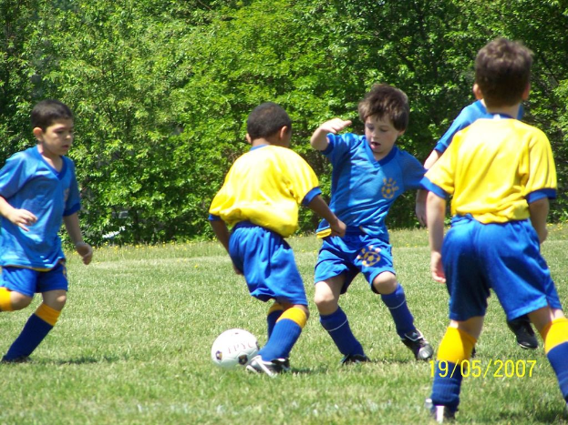 Kinder beim Fußballspielen auf einem grünen Feld mit Bäumen im Hintergrund und einem Datum in der rechten unteren Ecke.