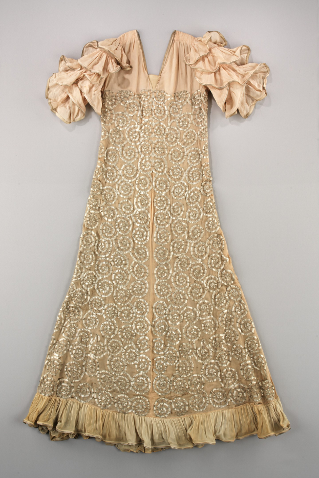 Ein cremefarbenes Kleid mit goldener Stickerei und Rüschenärmeln, das auf einer weißen Oberfläche präsentiert wird und mit detaillierten Designs und Mustern verziert ist.