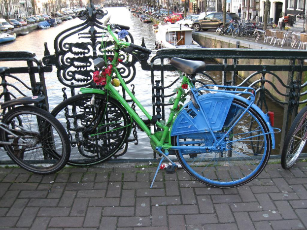 Fahrräder auf dem Boden geparkt mit einem Geländer dahinter, dahinter Wasser mit Booten; Autos und Gebäude säumen beide Seiten des Wassers, und es gibt Stühle und Tische in der oberen rechten Ecke.