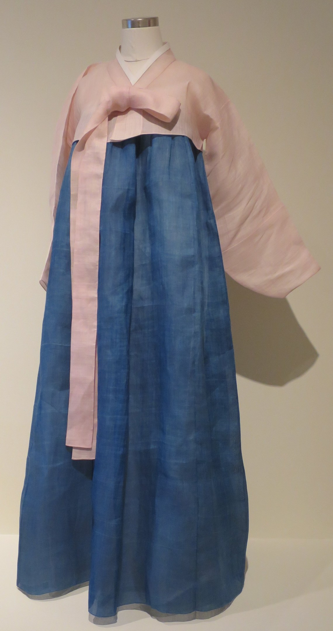 Eine Schaufensterpuppe in einem blauen und pinken hanbok, einem traditionellen koreanischen Kleid, das in einem Museum mit neutralem Hintergrund ausgestellt ist.