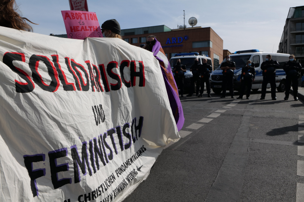 Eine Gruppe von Menschen marschiert die Straße entlang, hölt eine 'Solidarität und Feminismus'-Fahne, mit geparkten Fahrzeugen, Gebäuden, einer Schüsselantenne und einem klaren blauen Himmel im Hintergrund.