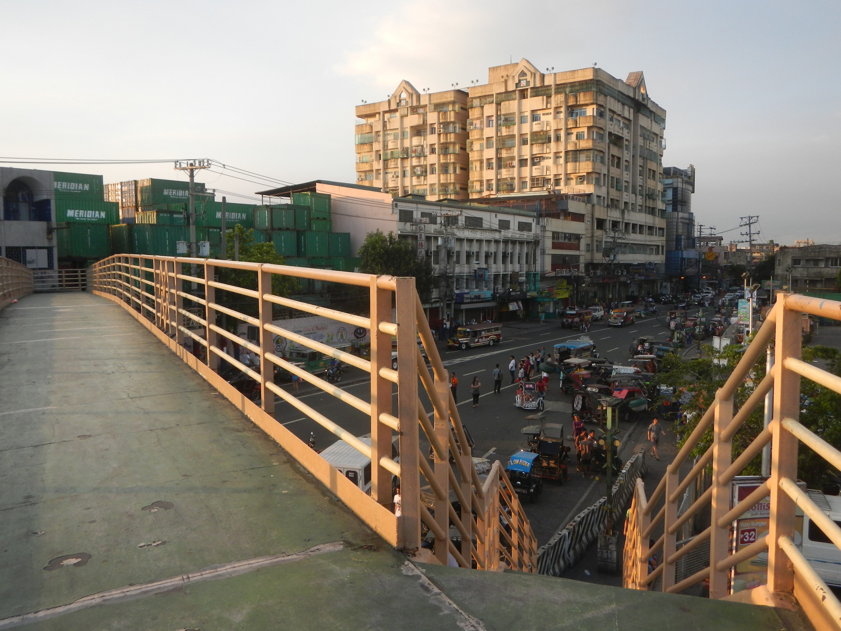 Fußgängerbrücke mit Geländern überspannt eine vielbefahrene Straße mit Fahrzeugen und Fußgängern, mit Gebäuden, Bäumen, Strommasten und einem bewölkten Himmel im Hintergrund.