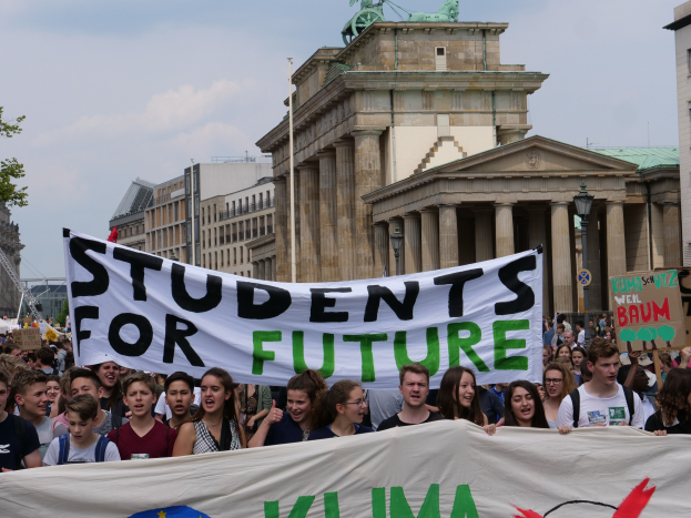 Gruppe von Studenten marschiert in Berlin mit einem buntfarbenen "Students for Future"-Plakat vor einer Kulisse aus Gebäuden, Bäumen und Himmel.
