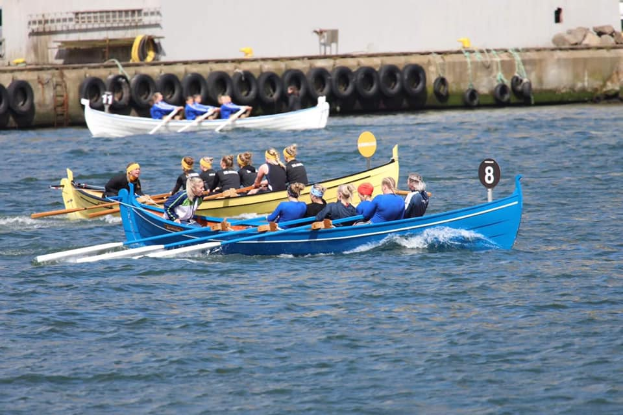 Eine Gruppe von Menschen in einem blauen und gelben Boot auf dem Wasser, mit Paddeln in der Hand, vor einer Wand mit Reifen und einem Gebäude im Hintergrund, was auf eine Regatta hindeutet.