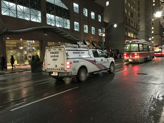 Ein Polizei-Lkw parkt nachts am Straßenrand, mit Fahrzeugen auf der Straße, Fußgängern auf dem Gehweg, Topfpflanzen, Straßenlaternen, Gebäuden mit Fenstern und einem bewölkten Himmel.