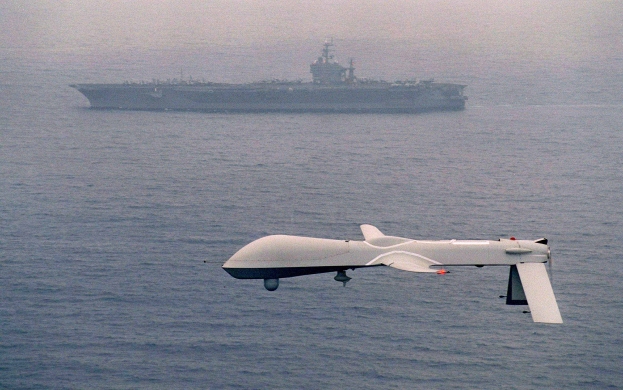 Weißes RQ-1 Predator-UAV fliegt über das Meer mit einem Schiff im Hintergrund.