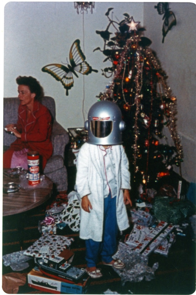 Ein Kind in einem Helm steht neben Geschenkverpackungen, Schachteln und einem Weihnachtsbaum auf dem Boden, während eine Frau in einem roten Kleid auf einem Sofa sitzt und eine Flasche sowie andere Gegenstände auf einem nahen Tisch stehen, mit zwei Schmetterlingsaufklebern an der weißen Wand im Hintergrund.