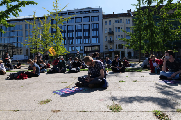 Eine Gruppe von Menschen sitzt vor einem Gebäude auf dem Boden während einer Demonstration in Berlin, einige tragen Masken und Habseligkeiten sind um sie herum verstreut, unter einem klaren blauen Himmel mit Bäumen und Gebäuden im Hintergrund.