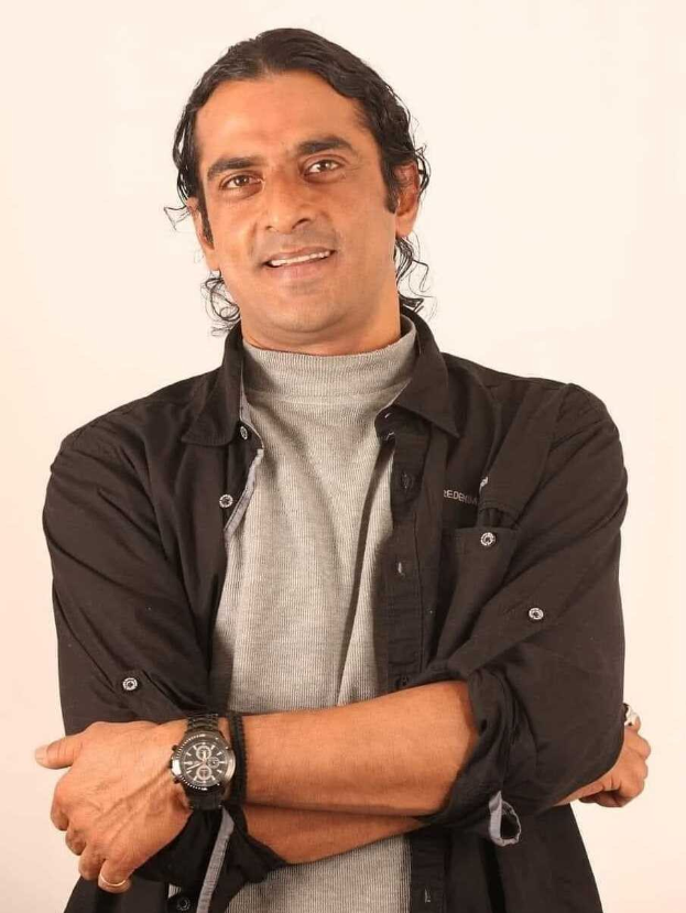 Ein Mann in einem schwarzen und grauen Outfit mit einer Uhr steht mit verschräckten Armen vor einem weißen Hintergrund und sieht nachdenklich aus.